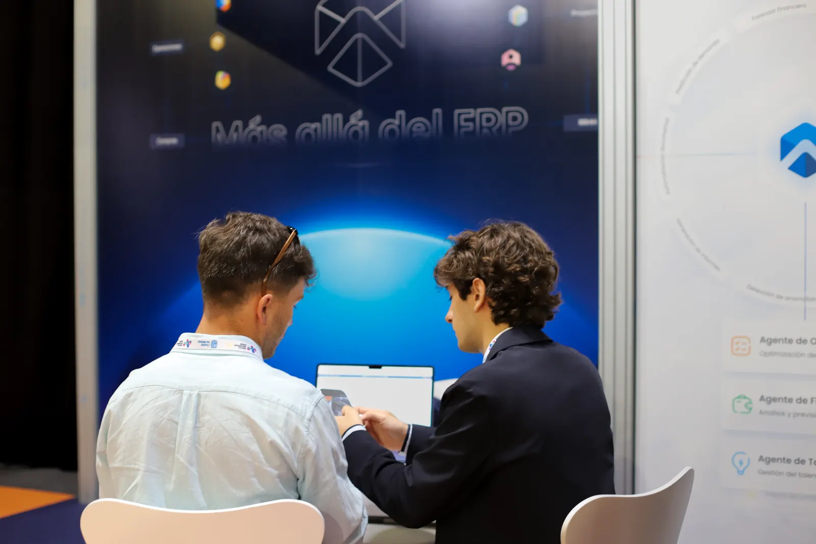 Two visitors over a laptop in front of the AI agents panel — IA integrada, no añadida